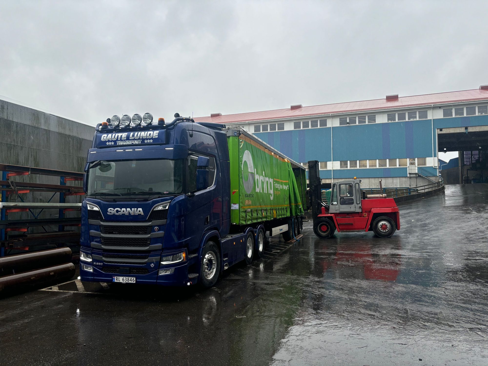 Mørkeblå trekkvogn med gardintrailer og en truck på et regnvått terminalområde.