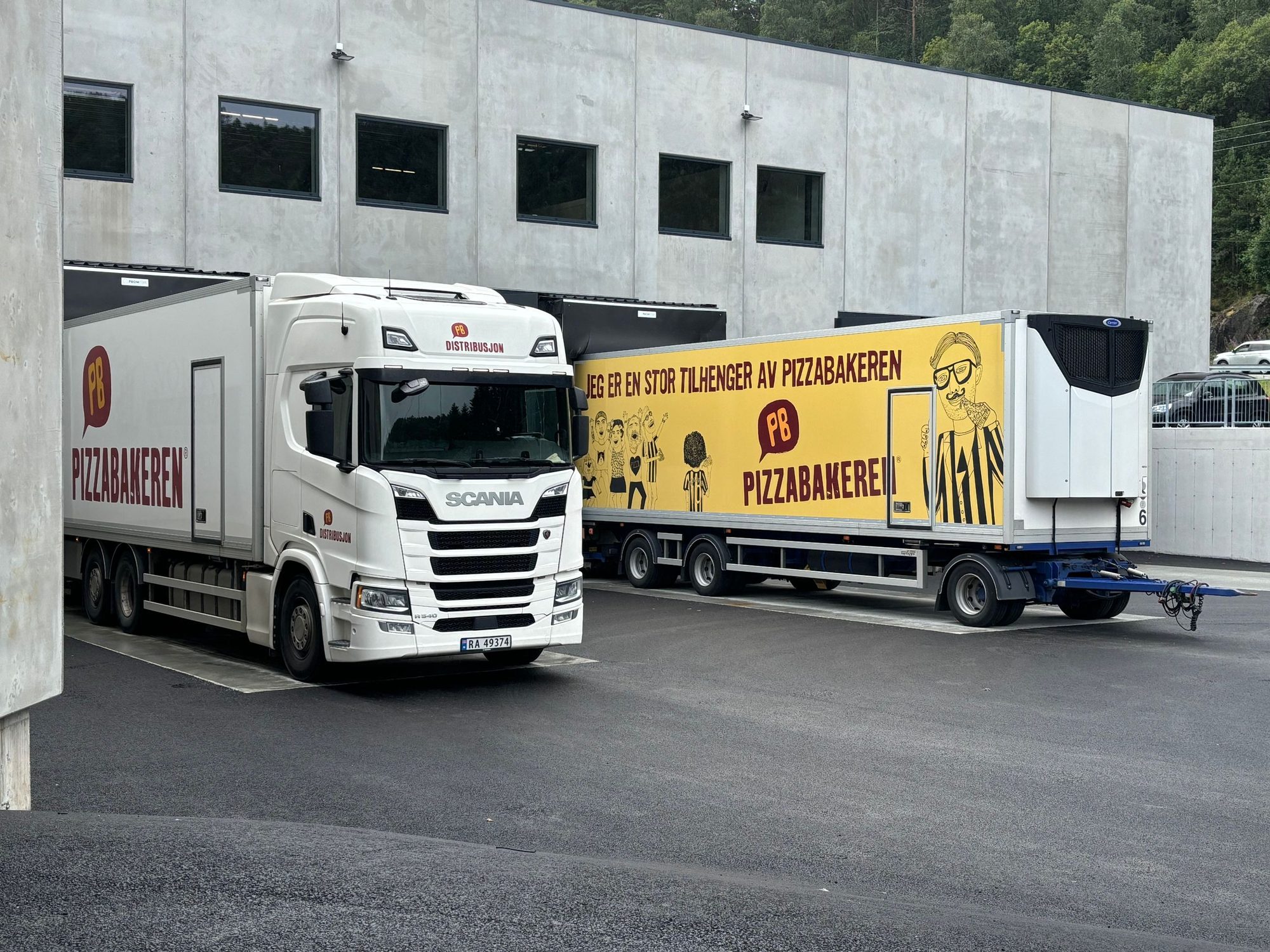 Distribusjonsbil med tilhenger parkert ved en lasterampe på et industribygg.